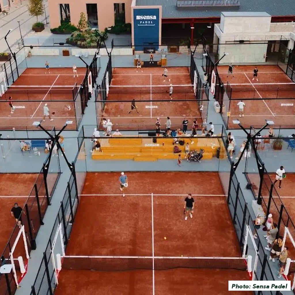 Aerial view of Sensa Padel Nashville at dusk, six clay courts busy with players, tiered seating in the middle and the club's sign on the building behind