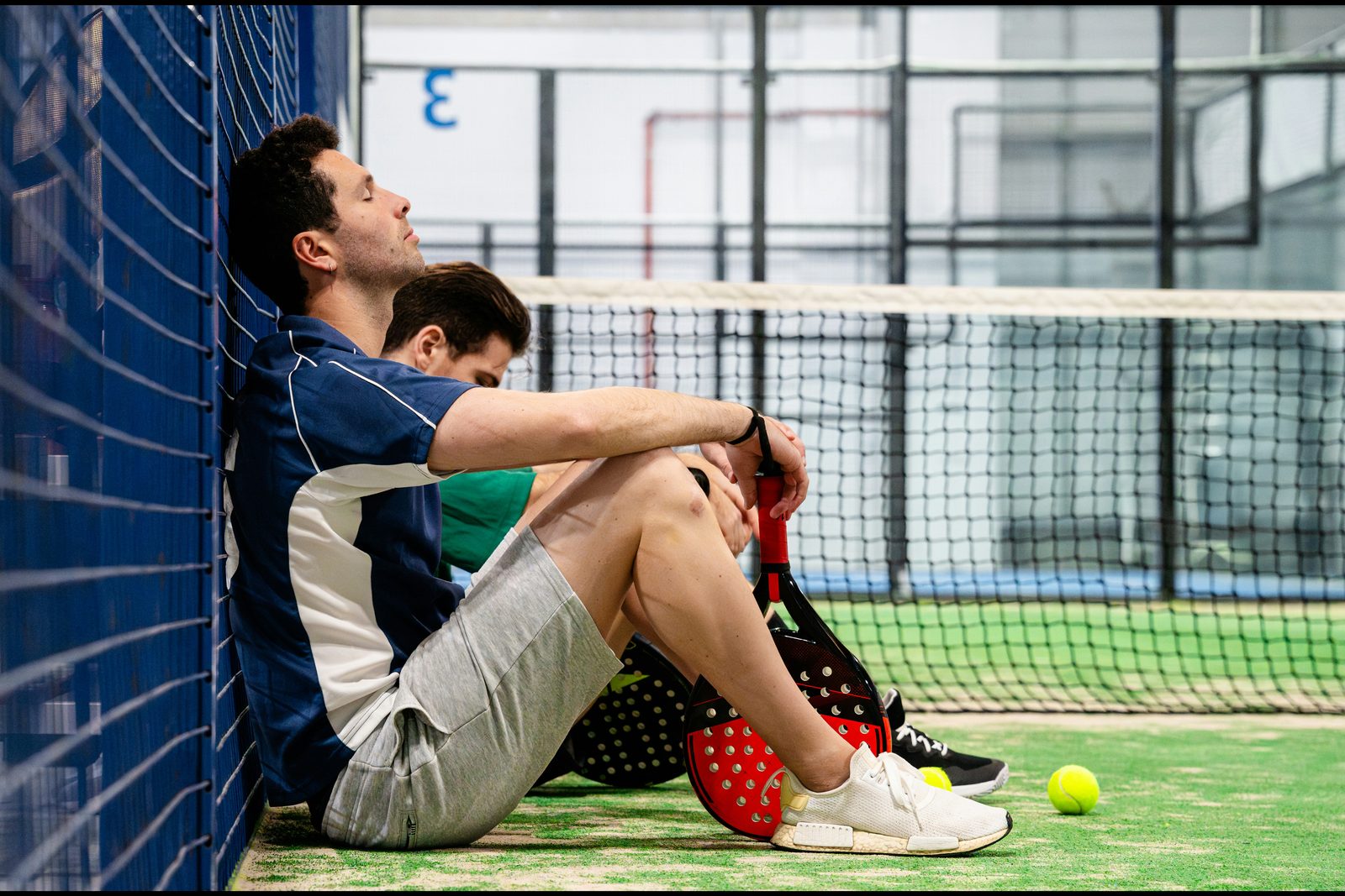Two padel players sitting against the court fence with their rackets, catching their breath after a match