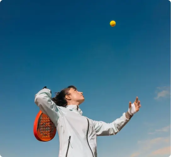 A woman in a white jacket tossing a padel ball into a clear blue sky, racket ready to serve.