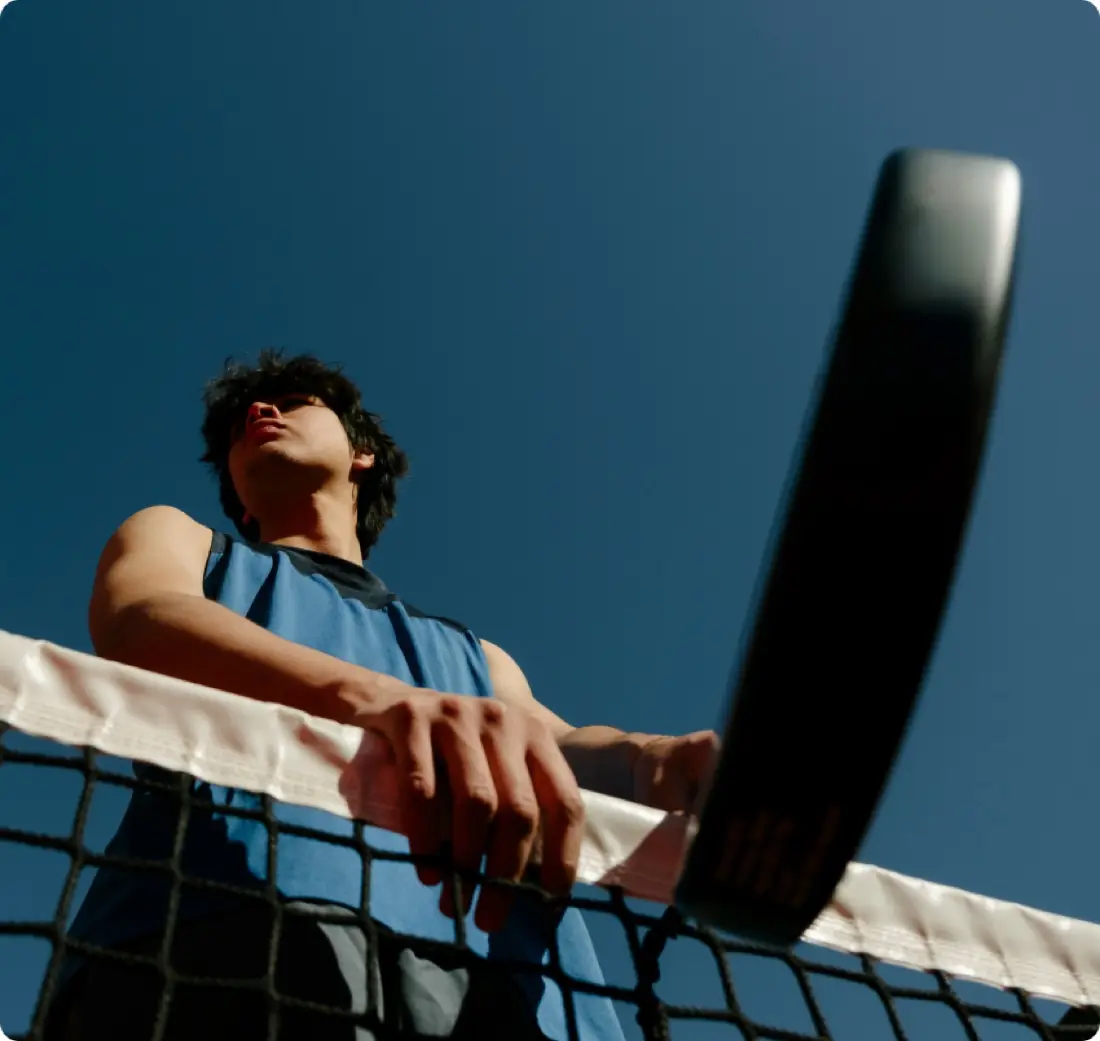 A padel player at the net, holding his paddle overhead, framed from below against a deep blue sky.