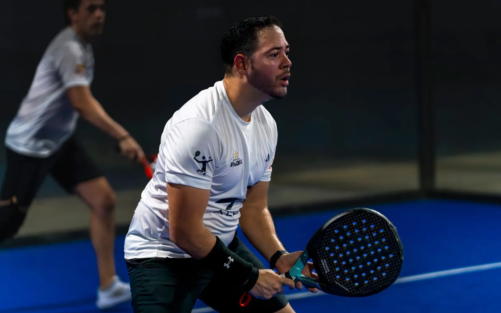 Two men playing padel doubles on an indoor court, the near player in a technical tee and shorts, ready at the net.